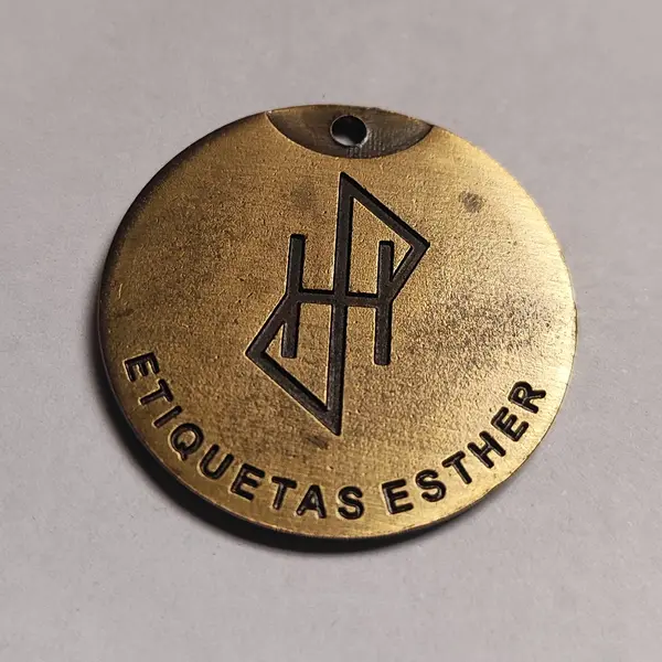 La misma medalla redonda de zamak en bronce tamboreado, en primer plano, con el monograma grabado en hueco
