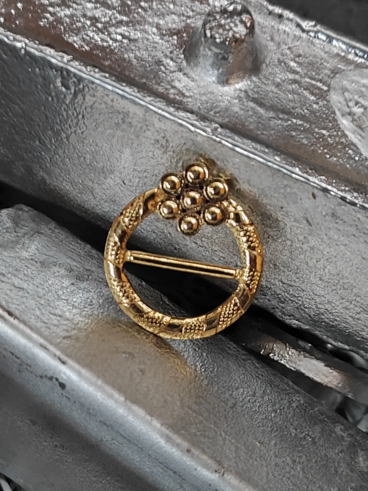 Hebilla de anillo en zamak con acabado dorado y un racimo de esferas en relieve, apoyada sobre lingotes de zamak